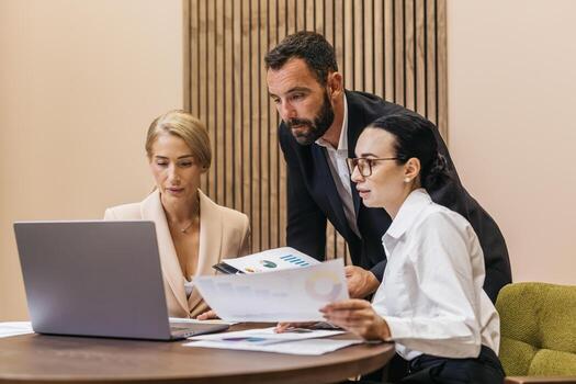 Business team of three professionals, two women and one man, collaborating at a modern office table, analyzing data on a laptop and discussing project strategies with printed reports photo
