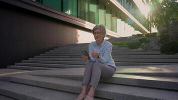 comovente tiro velho caucasiano empresária adulto meia idade mulher usando Móvel telefone ao ar livre digitando mensagem o negócio bate-papo olhando longe pensando resolver problema solução pensativo senhora sentar em cidade escadas video