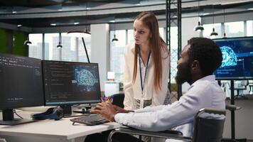 Junior dev and coworker in wheelchair developing software applications, optimizing code. Female technician and colleague with paraplegia in office using AI to solve bottlenecks, camera A video