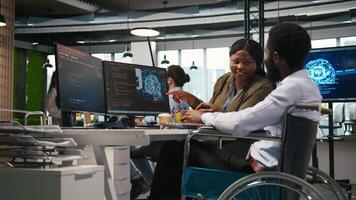 Smiling startup engineer and colleague with disability brainstorming, using machine learning tech. Happy african american man in wheelchair and coworker in office using AI predictive models, camera A video