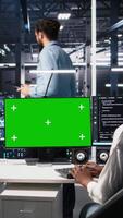 Vertical Female technician and coworker in data center review AI diagnostics on isolated screen PC. IT expert and colleague optimize artificial intelligence systems using chroma key computer, camera A video