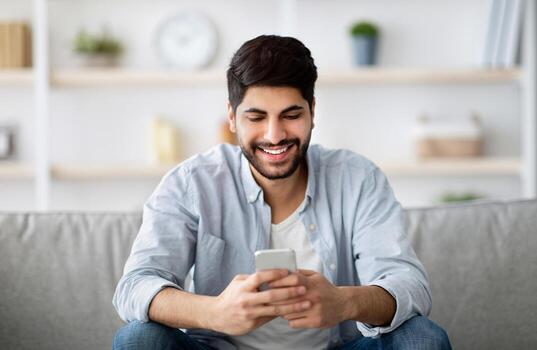 Handsome smiling arab man sitting with smartphone in hands and messaging with friends, relaxing with cellphone on couch, checking new app or browsing social networks, free space photo