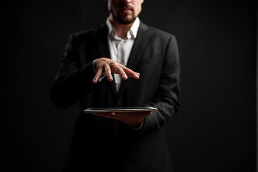 Man in formal attire presenting a tablet while gesturing with his hand in a dark studio setting photo