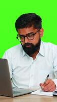 indio hombre se sienta a escritorio escritura ideas en cuaderno mientras trabajando en computadora portátil, demostración enfocar, planificación, multitarea árabe chico aislado en verde croma llave antecedentes expresando productividad pensamiento. video