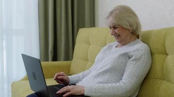 Happy retired woman using laptop computer, typing on keyboard and smiling while sitting on comfortable sofa at home, enjoying modern technology and online communication video