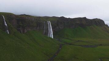 Seljalandsfoss waterfall cascades from a tall cliff in Iceland, with mist rising into an overcast sky. Visitors walk along a path at the base. video