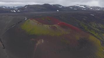 Aerial view of a volcanic crater in Iceland with red and green slopes, surrounded by dark terrain. Snow patches are visible on distant mountains. video