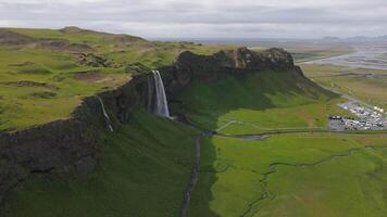 antenne visie van seljalandsfoss waterval in IJsland, trapsgewijs in een groen vallei met wandelen paden, een parkeren kavel, en ver weg bergen zichtbaar. video