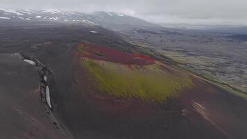 Aerial view of a volcanic crater in Iceland with red and green slopes, dark terrain, scattered snow patches, and a cloudy sky. Smooth camera motion. video
