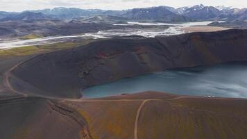 Aerial view of a volcanic crater lake with calm water, surrounded by dark soil, moss, and sparse vegetation. Snow capped mountains are visible in the background. video