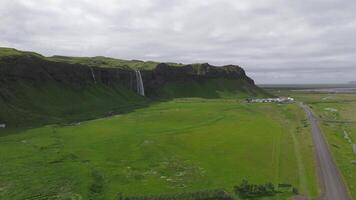 aéreo trasvolar de seljalandsfoss cascada en Islandia, demostración cascada agua, verde campos, caminando caminos, acantilados, y nublado cielo. video