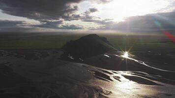 antenn se av en grön kulle omgiven förbi svart sand slätter med lindning strömmar. moln och solljus skapa dynamisk ljus och skugga effekter. video
