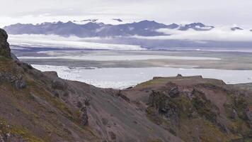 Aerial view of moss covered cliffs, glacial rivers, and a serene lagoon with icebergs. Distant mountains and clouds add depth to the tranquil scene. video