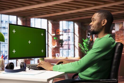 Cheerful man at home using green screen computer monitor to browse social media website, looking at memes. Smiling person in apartment looking at funny content online on chroma key PC display photo