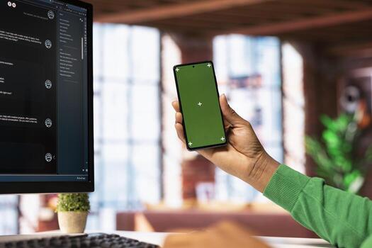 Man seated at desk using AI LLM chatbot software on PC and green screen phone in modern home office. Person holding mockup smartphone requesting help from large language model program photo