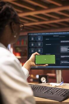 mujer sentado a escritorio utilizando ai llm larva del moscardón aplicación y aislado pantalla teléfono en moderno urbano hogar oficina, de cerca. africano americano persona usos grande idioma modelo programa en computadora y Bosquejo teléfono inteligente foto