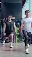 Two men execute synchronized dumbbell lunges in a well-lit gym environment, showcasing strength and coordination video