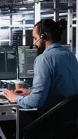 Vertical Engineer in data center wearing headset assisting users with AI enhanced troubleshooting. Server room employee managing support tickets using automated response systems, camera A video