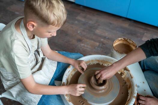 A blond boy learns pottery, sculpts clay on a pottery wheel and enjoys creative handicrafts in a studio under the guidance of a master photo