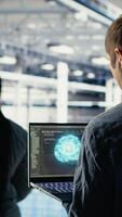 Vertical Data center system administrators investigate neural networks configuration, adjusting for optimal function. Server hub employees use laptop to check deep learning error reports, camera B video