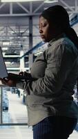 Vertical Data center admin investigates AI configurations using laptop, making adjustments to ensure optimal function. African american woman in server hub checks deep learning errors reports, camera A video