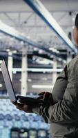 Vertical Data center admin investigates AI configurations using laptop, making adjustments to ensure optimal function. African american woman in server hub checks deep learning errors reports, camera B video