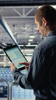 Vertical Server room IT expert swiping on tablet touchscreen to read data charts, monitoring equipment health. Admin reviews analysis metrics graphs on device to make sure data center meets goals, camera A video