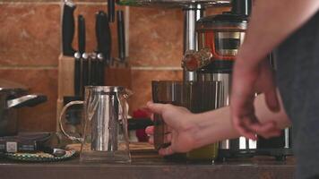 A close-up of a hand pouring fresh juice from a cold press juicer into a glass pitcher, with kitchen counters and various utensils in the background, showing the process of making healthy drinks. video