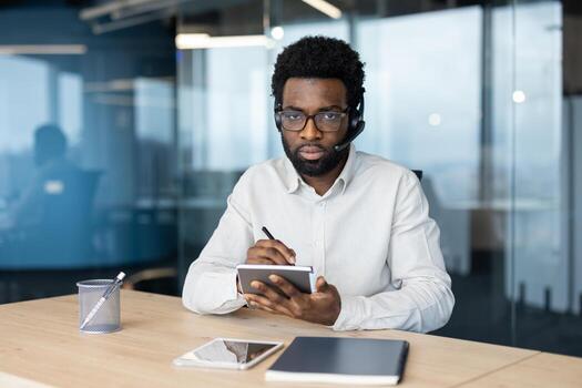 A businessperson with a headset working at a table, writing on a notepad, and using technology including a smartphone and tablet in a modern office environment. photo