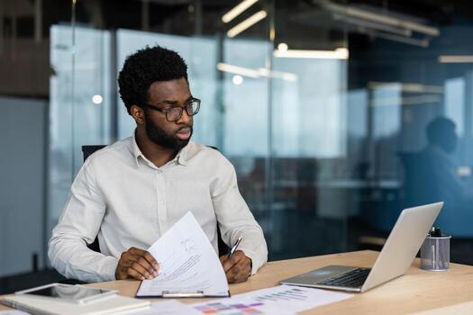 Young professional man engaged in reviewing documents while seated at a desk in a modern office environment. He displays focus and dedication, utilizing a laptop and papers to complete his tasks. photo