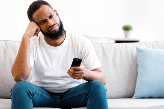 Boring Television Program. Bored African American Guy Watching TV Switching Channels With Remote Control Sitting On Sofa At Home. Discontented Viewer Watching Uninteresting Movie Concept. Copy Space photo