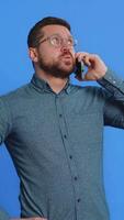 desinteresado hombre en lentes teniendo irritado aburrido hablar en teléfono inteligente chico cansado soñoliento persona de libre dedicación hablando en móvil teléfono con amigo, haciendo en línea conversacion aislado en azul antecedentes video