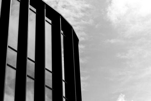 Black and white photo of a modern glass building with vertical lines and reflective windows, set against a cloudy sky. Suitable for illustrating architecture, urban design, or contemporary business concepts.