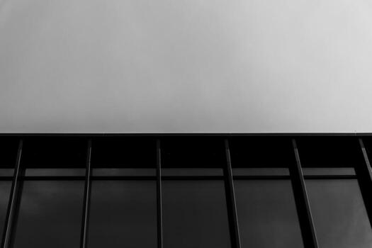 Black and white photo showing the upper section of a modern glass building with strong vertical lines and a clear sky above. Suitable for illustrating concepts of architecture, minimalism, and urban design in editorial or commercial projects.