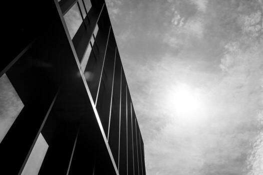 Black and white photo of a modern building facade with strong vertical lines and sunlight in a cloudy sky. Suitable for illustrating architecture, urban design, or minimalism concepts in editorial and commercial projects.