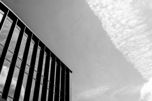Black and white photo of a modern building facade with vertical lines and glass windows against a sky with textured clouds. Suitable for illustrating architecture, urban design, or minimalism concepts in editorial or commercial projects.