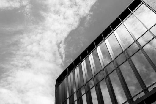 Black and white photo of a modern glass building facade reflecting clouds, captured from a low angle. Suitable for illustrating architecture, urban design, or business concepts in editorial and commercial projects.