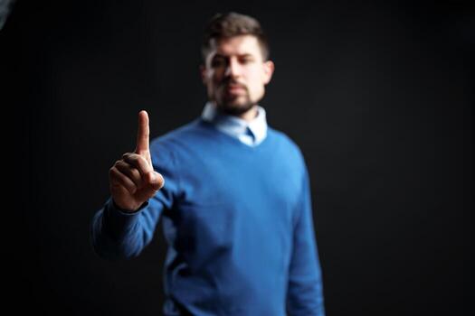 Man in blue sweater raises hand with index finger pointing up in dark setting to convey a message photo