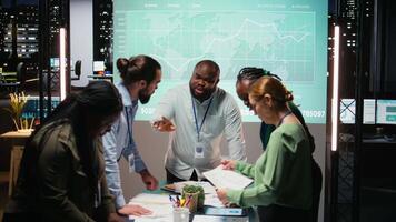 nacht tijd vergadering ontvouwt zich net zo een multi-etnisch team verzamelt in een strategie kamer, analyseren zichtbaar gegevens, statistieken en risico beoordeling bestanden naar rit project ontwikkeling voor efficiëntie. camera a. video