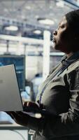Vertical Data center admin monitors deep learning process with laptop, reviewing operational metrics. African american woman using device to review neural networks reports during inspection, camera B video