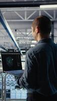 Vertical Software developer in data center checks AI dashboard using laptop during routine systems review. Server hub IT specialist tests artificial intelligence on notebook to check performance, camera A video