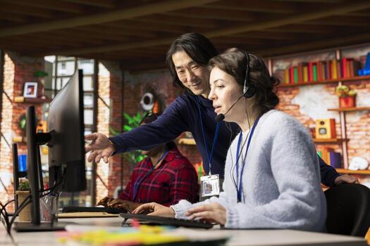 Smiling Asian call center manager evaluating company intern performance. Friendly experienced customer service department supervisor overseeing new employee technical onboarding photo