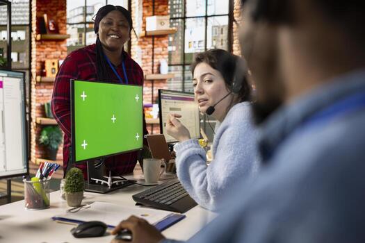 Call center agents laughing in office whit green screen mockup PC monitors, taking break. Customer support workspace colleagues having fun together at desks in front of chroma key computer display photo