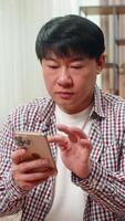 asiatico uomo su casa divano riempimenti in linea modulo su smartphone, accuratamente maschiatura schermo focalizzata grave sguardo. Cinese tipo Spettacoli diligente concentrazione preciso si sposta per evitare errori durante digitale applicazione video