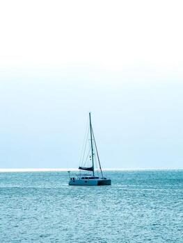 A low-angle shot captures a peaceful and expansive ocean scene. A white catamaran with its mast and rigging stands alone in the center, sailing calmly on the shimmering blue water. The horizon is a photo