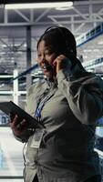 Vertical Data center system administrator uses AI on tablet to help customers resolve website hosting problems. African american woman in server farm helps users keep online services running smoothly, camera A video