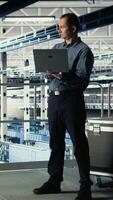 Vertical Data center admin on industrial platform checks AI dashboard using laptop. Man in server farm tests artificial intelligence tools on notebook to ensure reliable performance, camera A video