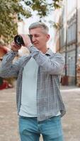 de edad mediana hombre turista de viaje en fines de semana explorador pueblo mirando alrededor ciudad, haciendo foto imágenes en cámara al aire libre. fotógrafo maduro chico viajero en urbano pueblo calle. vertical. estilos de vida video