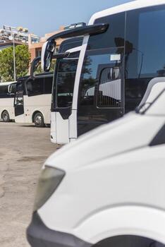 A variety of buses in different colors are parked at a travel terminal on a sunny day. The vehicles are lined up neatly, with a few large buses and small vans in view photo