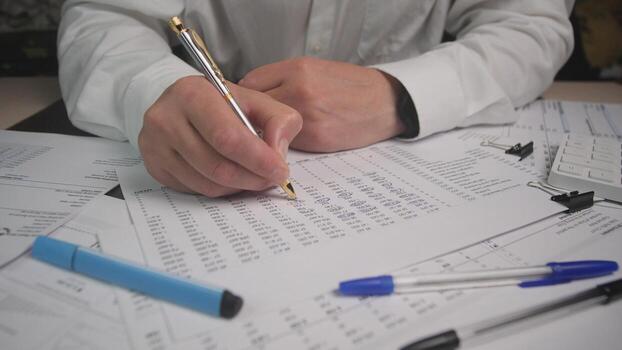 Individual analyzing financial sheets and making notes in an office setting, surrounded by various writing instruments and paperwork, emphasizing organization and focus. photo